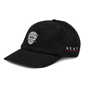 Op de afbeelding: Zwarte corduroy baseballcap met een wit geborduurd hockeymasker op de voorkant. Het woord "HEAT" is in het wit geborduurd met een rode onderstreping aan de zijkant. De cap heeft een gebogen klep.