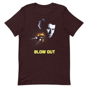 May include: A maroon t-shirt with a graphic of a man's face and a car in a target scope. The text "BLOW OUT" is printed in yellow below the graphic.