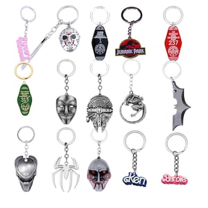 Op de afbeelding: Een set van 15 zilverkleurige sleutelhangers met verschillende film- en popcultuurpersonages. De sleutelhangers bevatten personages uit Jurassic Park, The Overlook Hotel, The Great Northern Hotel, Friday the 13th, Alien, Batman, Predator, Spider-Man, en meer.