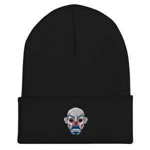 K&ouml;nnte beinhalten: Schwarze M&uuml;tze mit einem gestickten Design eines Clowngesichts. Das Clowngesicht hat rote Augen, ein wei&szlig;es Gesicht und einen blau-roten Mund. Die M&uuml;tze ist aus einem weichen, gestrickten Material gefertigt.