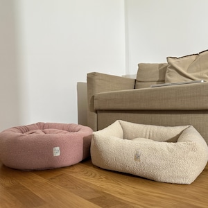 Puede incluir: Dos camas para perros, una rosa y otra beige, están en un suelo de madera. Las camas están hechas de un material suave y esponjoso y tienen un borde elevado. La cama rosa es redonda y la cama beige es rectangular.