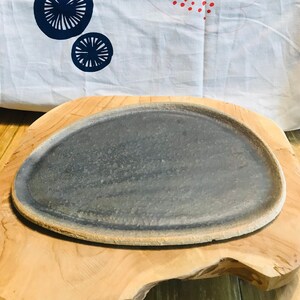 Peut inclure: Une assiette ovale en céramique gris foncé avec un bord légèrement surélevé repose sur une surface en bois brun clair. L'assiette a une surface texturée et un léger éclat.