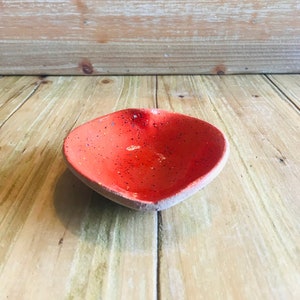 Puede incluir: Un pequeño plato en forma de corazón hecho de cerámica roja moteada. El plato está sobre una superficie de madera.