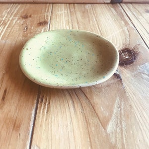 Peut inclure: Un petit plat en céramique fait à la main, avec une base vert clair et des accents mouchetés bleus et noirs. Le plat de forme ovale repose sur une surface en bois, mettant en valeur son design artisanal unique.