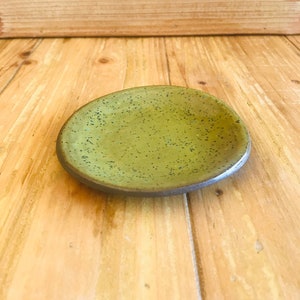 May include: A small, green ceramic dish with a speckled glaze. The dish is slightly rounded and has a dark brown rim.