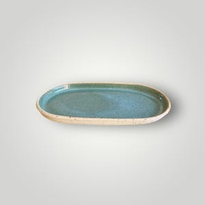 May include: An oval ceramic serving platter with a speckled teal interior and a light beige rim. The handmade pottery piece has a rustic, organic aesthetic, perfect for displaying food or decorative items.