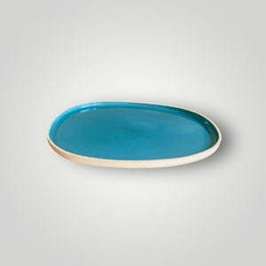 Peut inclure: Un plat de service ovale en céramique, fait à la main. L'intérieur est turquoise vif, contrastant avec le bord blanc cassé moucheté. Idéal pour servir des hors-d'œuvre ou pour présenter des objets décoratifs.