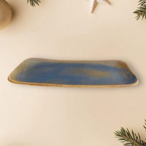 Könnte beinhalten: Ein rechteckiges blaues und goldenes Keramik-Serviertablett mit leicht erhöhtem Rand. Das Tablett befindet sich auf einem hellbeigen Hintergrund mit goldenen Weihnachtsornamenten und Kiefernästen.