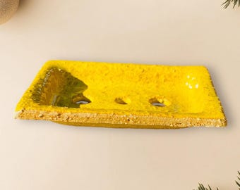 Puede incluir: Un jabonera de cerámica amarilla con tres agujeros de drenaje. La jabonera tiene una superficie texturizada y una forma ligeramente irregular.