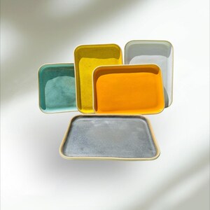 Puede incluir: Conjunto de bandejas rectangulares para servir en varios colores: turquesa, amarillo, naranja, gris y gris oscuro. Cada bandeja tiene un borde de color claro, creando contraste con los vibrantes interiores. Las bandejas están dispuestas de forma atractiva.
