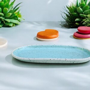 Peut inclure: Un plateau ovale en céramique bleu clair avec des points blancs. Le plateau est vide et posé sur une surface blanche.