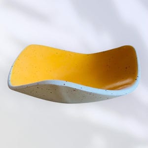 May include: A ceramic dish with a wavy design. The interior is a vibrant yellow with small dark speckles, while the exterior is a speckled grey with a light blue rim. The dish has a smooth, glossy finish.