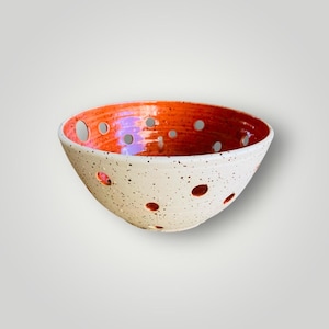 Op de afbeelding: Een gespikkelde witte keramische kom met een oranje binnenkant. De kom heeft verschillende ronde gaten en een breed, open ontwerp. De binnenkant is fel oranje van kleur, wat contrasteert met de gespikkelde witte buitenkant. Deze handgemaakte kom is ideaal om te serveren of tentoon te stellen.
