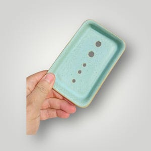 Puede incluir: Una jabonera rectangular de cerámica en color verde menta claro con un acabado moteado. El recipiente cuenta con un borde elevado y una serie de cinco orificios de drenaje circulares dispuestos en línea en el centro para el paso del agua.