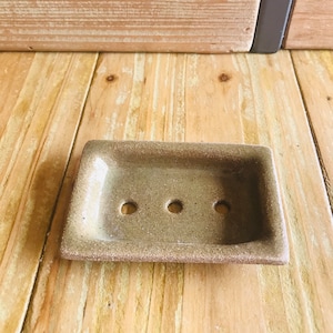 Puede incluir: Un pequeño jabonera rectangular de cerámica marrón moteada con tres agujeros de drenaje. La jabonera está sobre una superficie de madera.