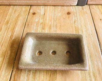 Puede incluir: Un pequeño jabonera rectangular de cerámica marrón moteada con tres agujeros de drenaje. La jabonera está sobre una superficie de madera.