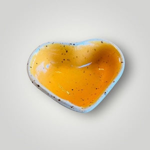 May include: A heart-shaped ceramic dish with a vibrant yellow interior and speckled detailing. The rim is a light blue color with a speckled gray border. This decorative dish is perfect for holding small items.