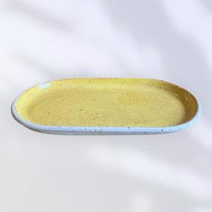 May include: A handmade, oval ceramic serving platter. The interior is a speckled yellow, while the rim is a speckled white. The platter is ideal for serving appetizers or small dishes. The dimensions are approximately 30 cm long and 15 cm wide.