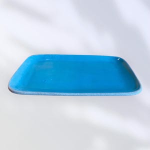 Può includere: Un vassoio rettangolare in ceramica turchese con angoli arrotondati. Il vassoio ha una finitura liscia e lucida e un bordo azzurro. Il design è semplice e moderno, adatto per servire o esporre.