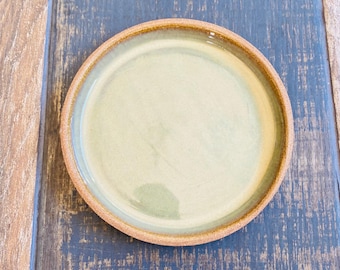 Może przedstawiać: Okrągły ceramiczny talerz z jasnozielonym środkiem i brązową obwódką. Talerz leży na niebiesko-brązowej drewnianej powierzchni. Prosty, ale elegancki element zastawy stołowej.