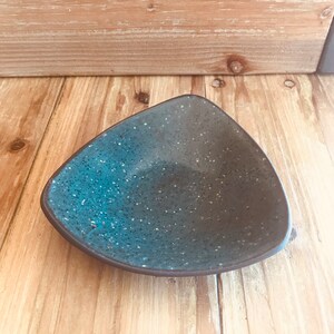 May include: A triangular ceramic dish with a speckled blue glaze. The dish is perfect for serving small snacks or dips.