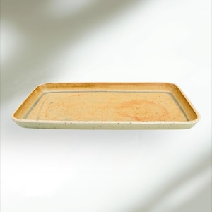 Puede incluir: Bandeja rectangular de cerámica con borde verde claro moteado e interior beige. Una fina línea azul bordea el interior. Ideal para servir alimentos o exhibir objetos decorativos.
