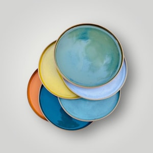 Pode incluir: Uma pilha de seis pratos redondos de cerâmica com uma variedade de cores vibrantes, incluindo laranja, amarelo, azul-petróleo, azul-claro e um verde-azulado manchado. Cada prato tem uma superfície lisa e vidrada com uma borda de tom neutro.