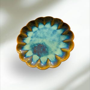 Op de afbeelding: Een keramische schaal met een geschulpte rand. De schaal heeft een blauw en groen midden met een bruine en oranje rand. De schaal heeft een glanzende afwerking en is waarschijnlijk handgemaakt. De schaal is rond en heeft een bloemachtige vorm.