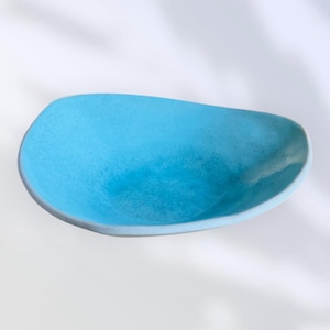 Puede incluir: Un cuenco de cerámica hecho a mano, de forma ovalada, en un vibrante color turquesa. El cuenco tiene una superficie ligeramente texturizada y un borde liso de color gris claro. Este cuenco decorativo es perfecto para servir aperitivos o como pieza decorativa.