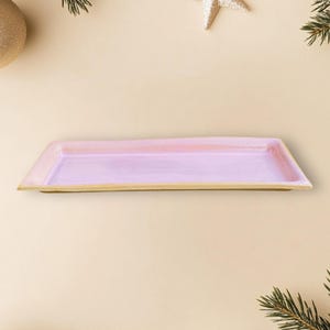 Peut inclure: Un plateau rectangulaire en céramique de couleur lilas clair avec un bord légèrement surélevé. Le plateau est sur un fond beige avec des décorations de Noël dorées et des branches de pin.