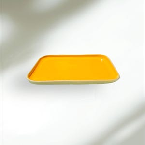 Puede incluir: Una bandeja rectangular para servir con un interior amarillo brillante y un borde verde claro. La bandeja tiene un acabado brillante y un diseño simple y moderno. Perfecta para servir aperitivos o exhibir objetos decorativos.
