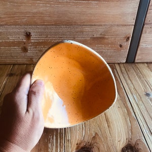 Puede incluir: Un cuenco de cerámica naranja triangular con un esmalte moteado. El cuenco se sostiene en una mano y descansa sobre una superficie de madera.
