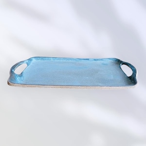 Peut inclure: Un plateau de service rectangulaire avec un glaçage bleu clair et deux découpes pour les poignées. Le plateau a une surface texturée et un bord naturel non émaillé. Le design est simple et élégant, adapté au service ou à l'exposition.