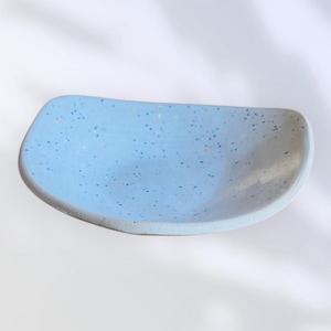 May include: A light blue ceramic dish with a speckled pattern. The dish has a curved rectangular shape with rounded edges. The speckled pattern includes small dots of various colors, adding visual interest to the handmade pottery.
