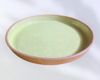 May include: A round, shallow ceramic bowl with a light green interior and a reddish-brown exterior. The rim of the bowl has a slightly raised edge with a pinkish hue. The bowl is filled with a light green liquid.