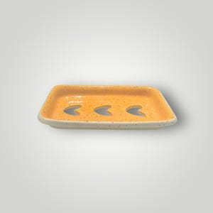 Peut inclure: Un porte-savon rectangulaire en céramique d'une teinte orange vif avec une finition mouchetée. L'objet présente trois découpes de drainage en forme de croissant au centre, un rebord surélevé et un vernis lisse et brillant sur toute la surface.
