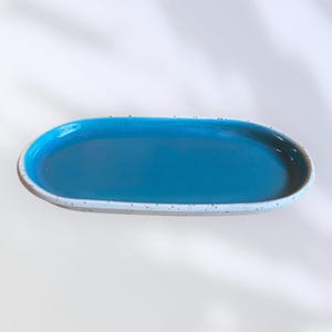 Peut inclure: Un plat de service ovale en céramique. L'intérieur est bleu turquoise vif, contrastant avec le bord blanc cassé moucheté. Le plat a une finition lisse et brillante, suggérant qu'il est conçu pour la présentation des aliments.
