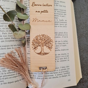 Puede incluir: Un marcap&aacute;ginas de madera con un dise&ntilde;o de &aacute;rbol grabado con l&aacute;ser y el texto "Bonne lecture ma petite Mamie". El marcap&aacute;ginas tiene un borla marr&oacute;n.