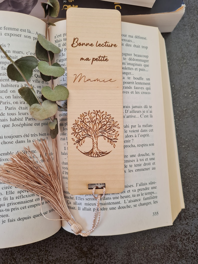 Puede incluir: Un marcap&aacute;ginas de madera con un dise&ntilde;o de &aacute;rbol grabado con l&aacute;ser y el texto "Bonne lecture ma petite Mamie". El marcap&aacute;ginas tiene un borla marr&oacute;n.
