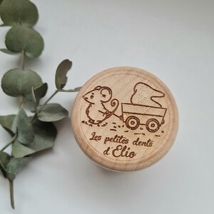 K&ouml;nnte beinhalten: Eine Holzkiste mit einer Maus, die einen Wagen mit einem Zahn darin schiebt. Die Kiste ist mit dem Text "Les petites dents d'Elio" graviert.