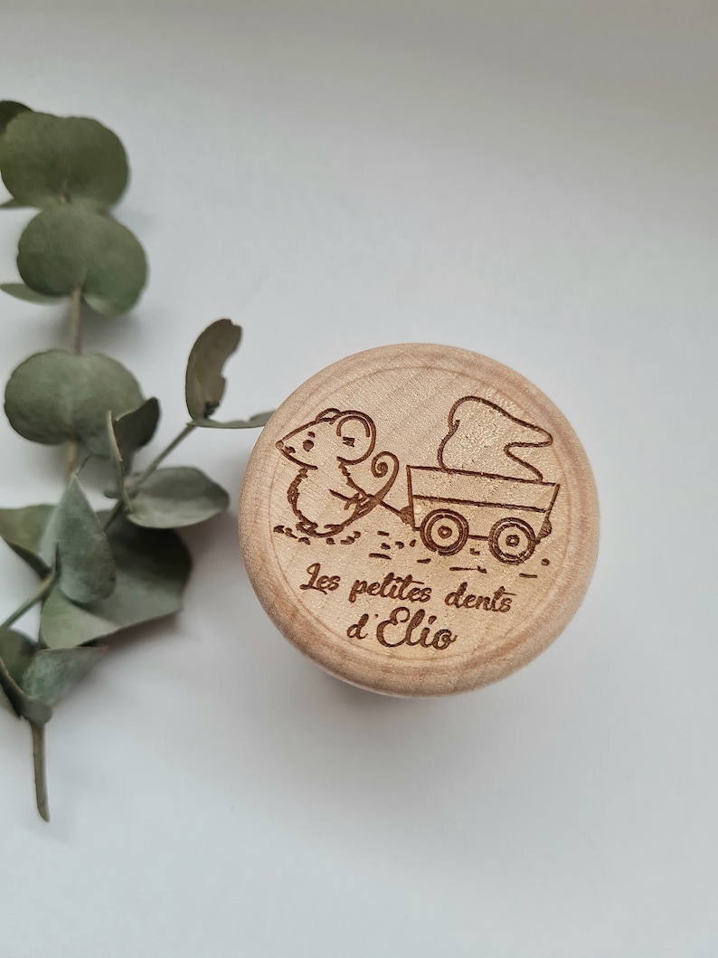 K&ouml;nnte beinhalten: Eine Holzkiste mit einer Maus, die einen Wagen mit einem Zahn darin schiebt. Die Kiste ist mit dem Text "Les petites dents d'Elio" graviert.
