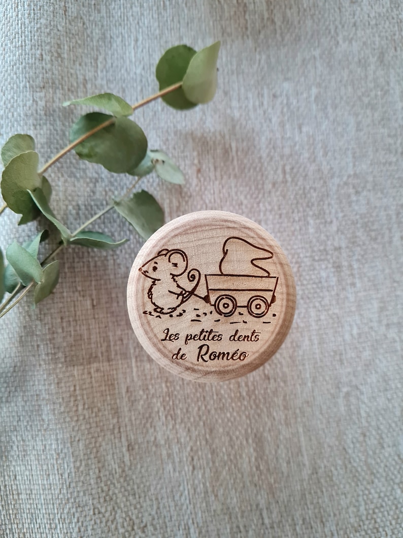K&ouml;nnte beinhalten: Eine Holzkiste mit einer Maus-Illustration, die einen Wagen mit einem Zahn darin schiebt. Die Kiste ist mit dem Text "Les petites dents de Romeo" graviert.