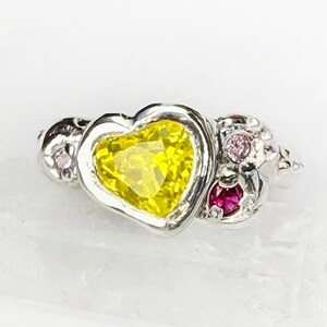 May include: A silver ring featuring a heart-shaped yellow gemstone as the centerpiece. The ring is adorned with smaller pink and red gemstones on either side of the heart, creating a delicate and feminine design. The ring is set on a white surface.