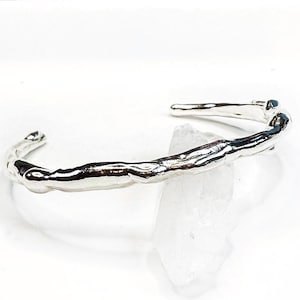 May include: A silver-colored cuff bracelet with a textured, organic design. The bracelet has an open design and appears to be made of metal. The surface reflects light, highlighting the irregular shape and texture of the jewelry.