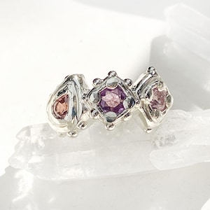 May include: A silver ring with three gemstones. The gemstones are pink, purple, and a darker pink. The ring is set on a white background.