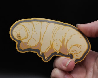 May include: A yellow and brown sticker of a tardigrade, also known as a water bear, on a black background.