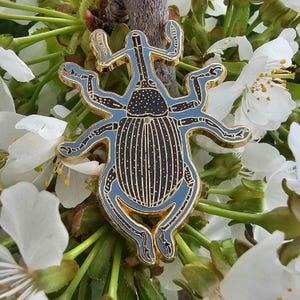 May include: Gold-colored pin of a blue and black weevil with intricate details. The pin is set against a background of white cherry blossoms.