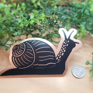 May include: Black and white snail sticker on a copper background. The snail is in a curled position with its shell facing the left side of the image. The sticker is approximately the size of a UK 20 pence coin.