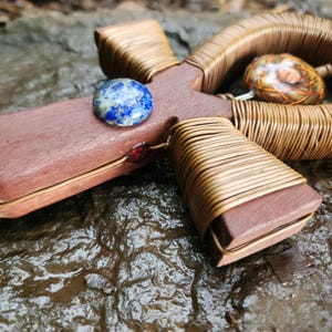 May include: A handcrafted wooden cross pendant with a lapis lazuli stone and wire wrapping. The cross features a brown wooden base, wrapped with gold-colored wire. A blue and white stone is set on the cross, with a smaller stone on the side.