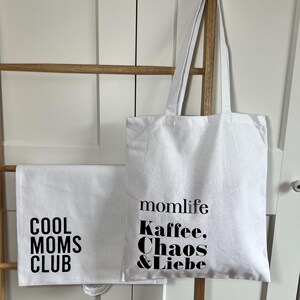 May include: White tote bag with black text that reads "momlife Kaffee, Chaos & Liebe" and a white tote bag with black text that reads "Cool Moms Club".